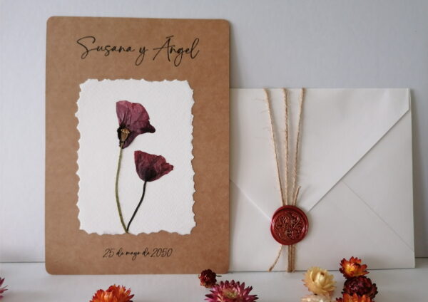 En esta imagen se ve una invitación de boda botánica. El fondo es papel de 300gr blanco roto con textura. Las medidas son 14,8x21cm y sobre este fondo hay un recuadro de papel Kraft de 300gr sobre el que hay pegadas dos amapolas que han sido prensadas y secadas por mi. En la parte superior están impresos los nombres de los novios y en la parte inferior la fecha. Los datos restantes están en el reverso de la tarjeta. También se ve un sobre de papel Kraft adornado con una cuerda y un sello de lacre color rojo.